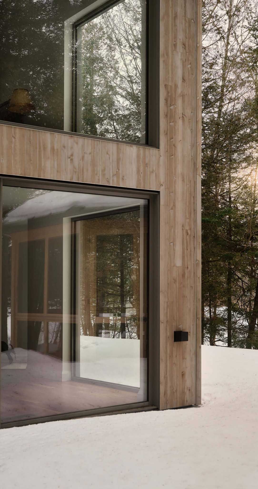 Modern cabin exterior with large windows, snow on ground, and forest in background.