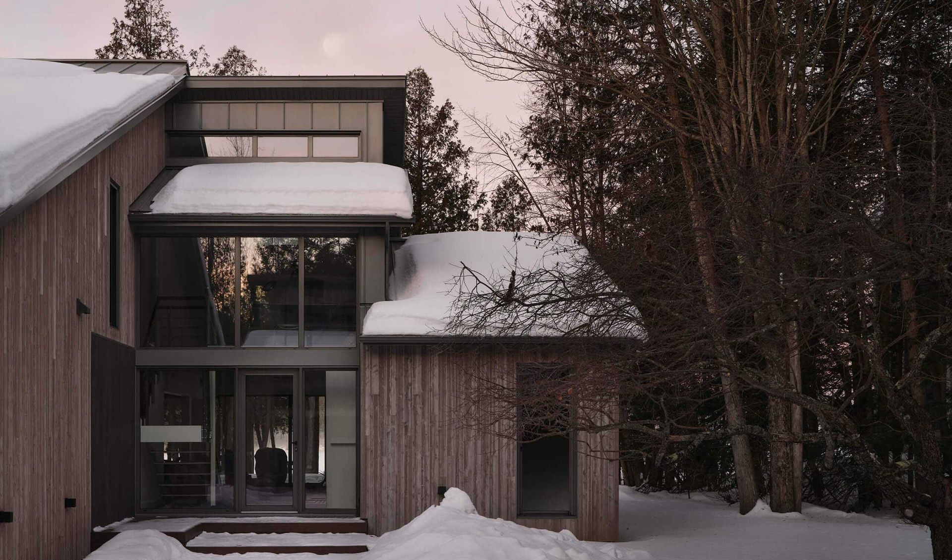 Maison moderne aux murs lambrissés, recouverte de neige, jouxtant une forêt enneigée en hiver.