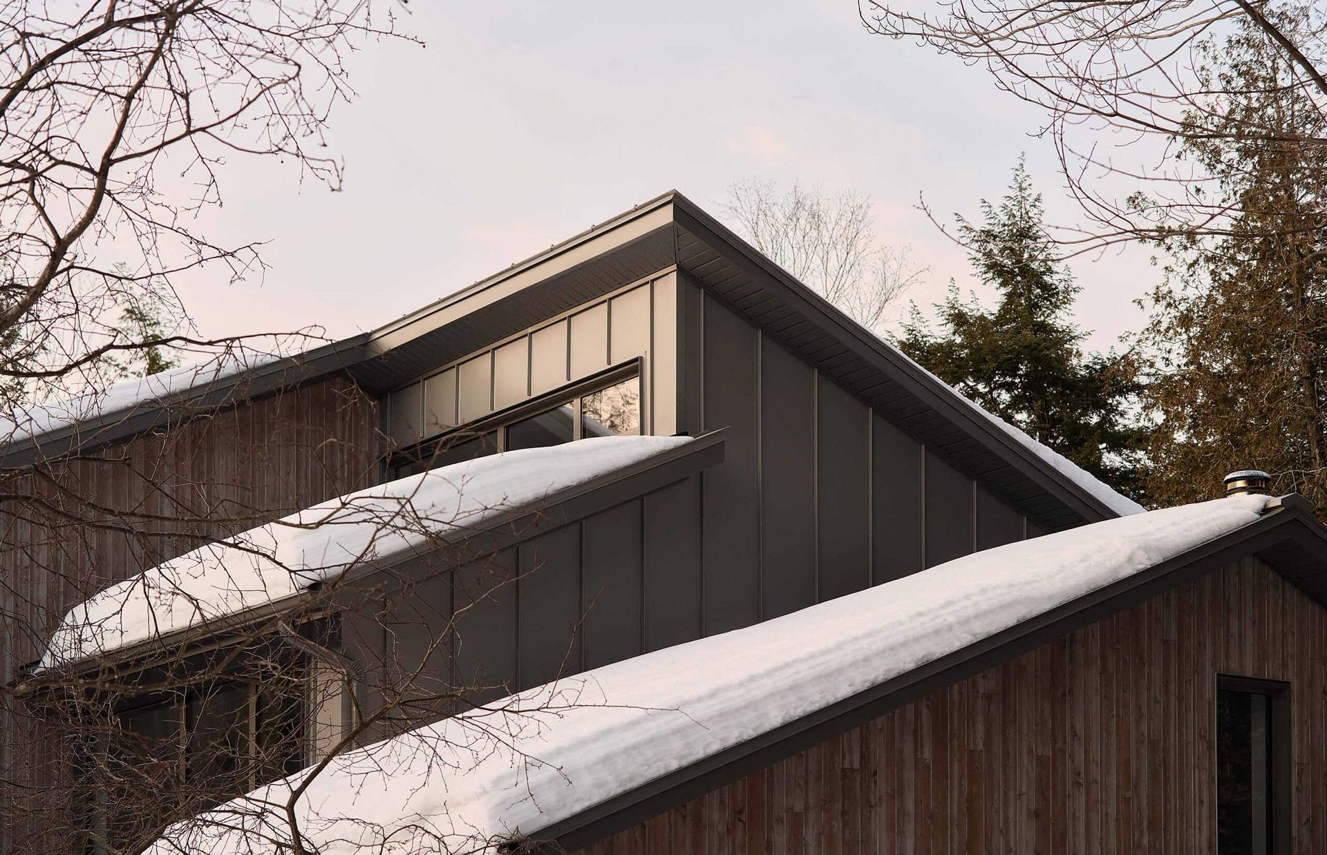 Ext&eacute;rieur d'une maison avec toit enneig&eacute;, bardage en bois et arbres dans un d&eacute;cor hivernal.