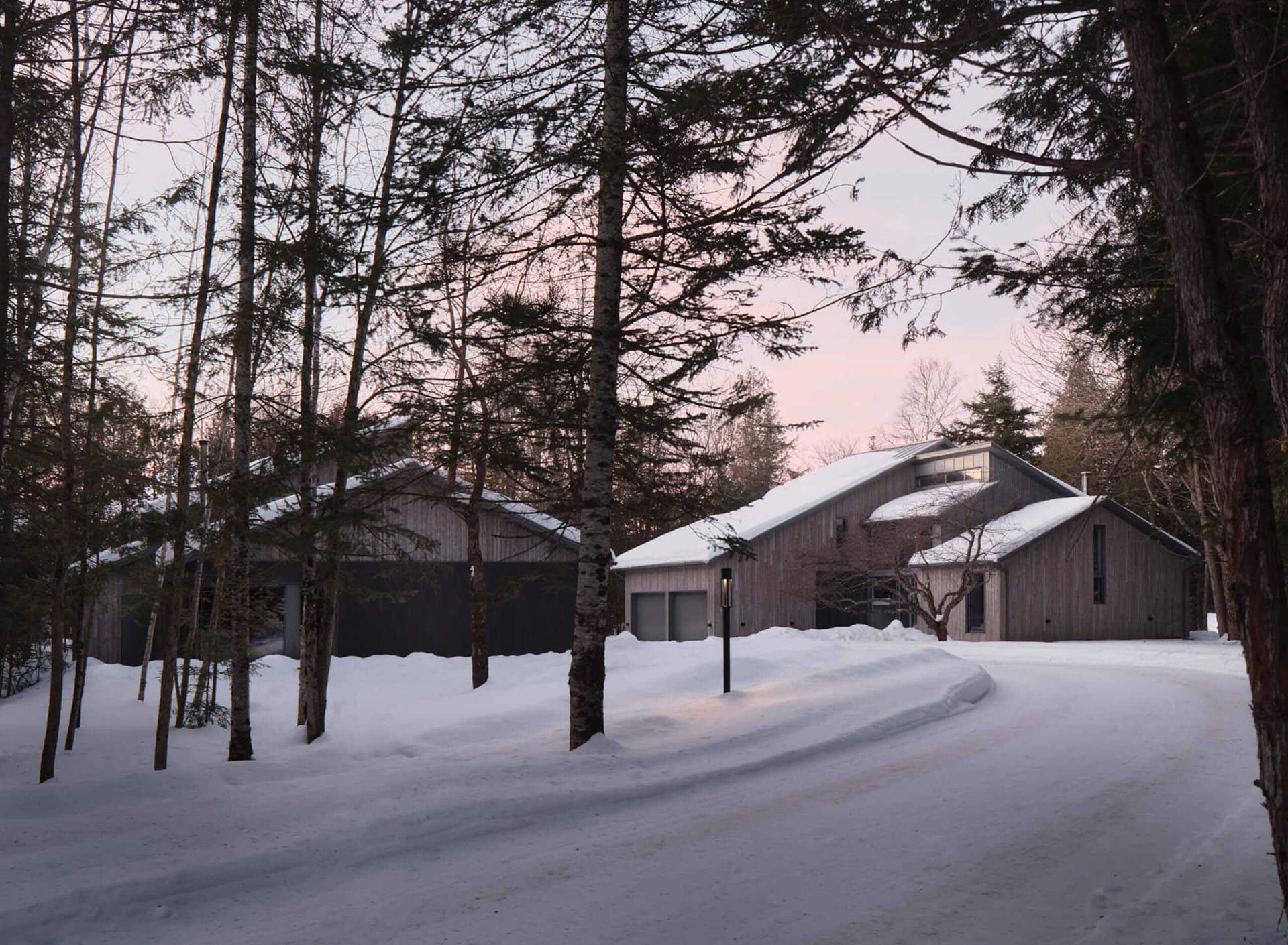 Un chemin enneig&eacute; m&egrave;ne &agrave; une maison et un garage dans un d&eacute;cor de for&ecirc;t hivernale ; ciel cr&eacute;pusculaire.