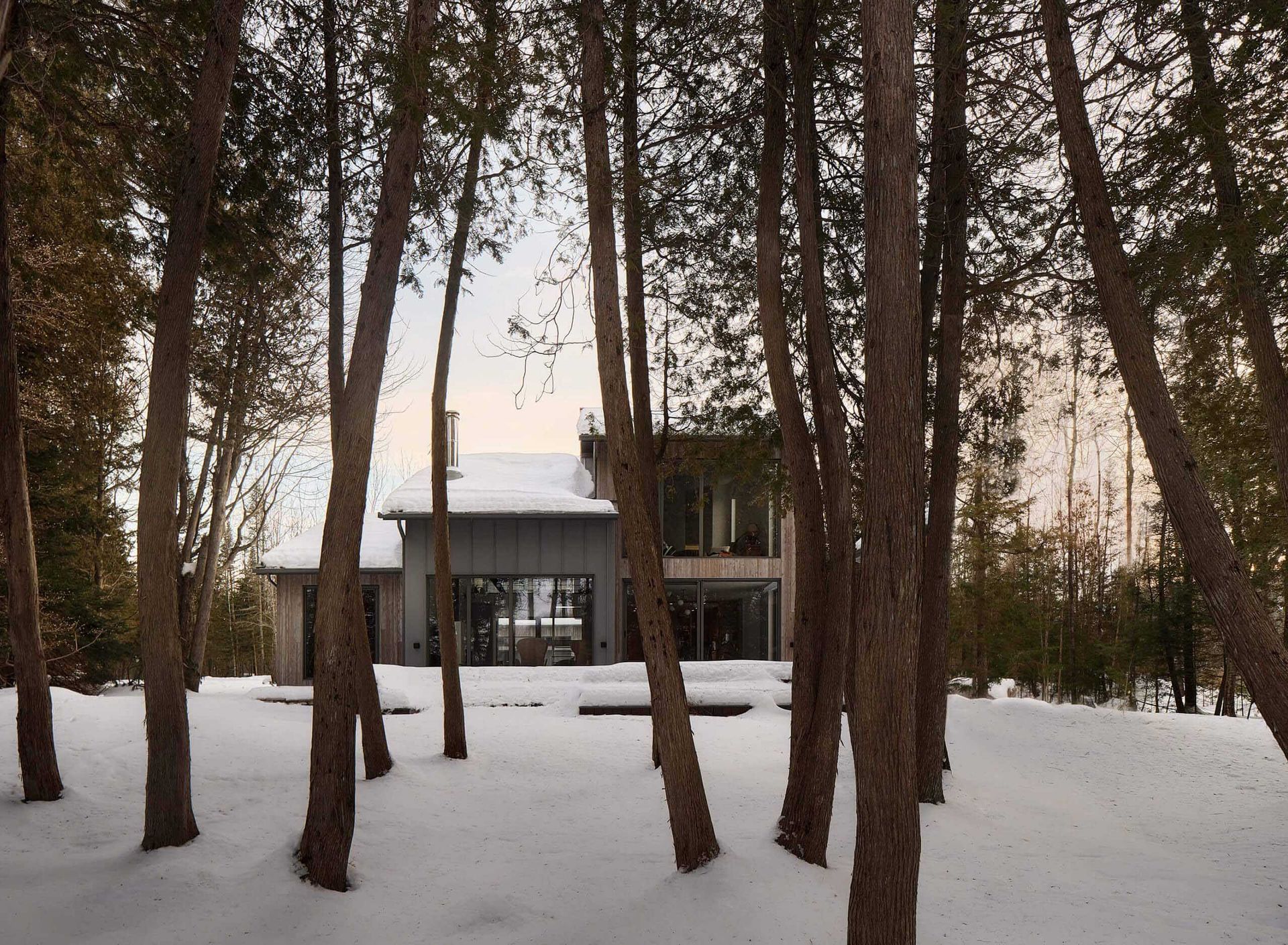 Maison nich&eacute;e dans une for&ecirc;t enneig&eacute;e, vue &agrave; travers les troncs d'arbres ; ciel couvert.