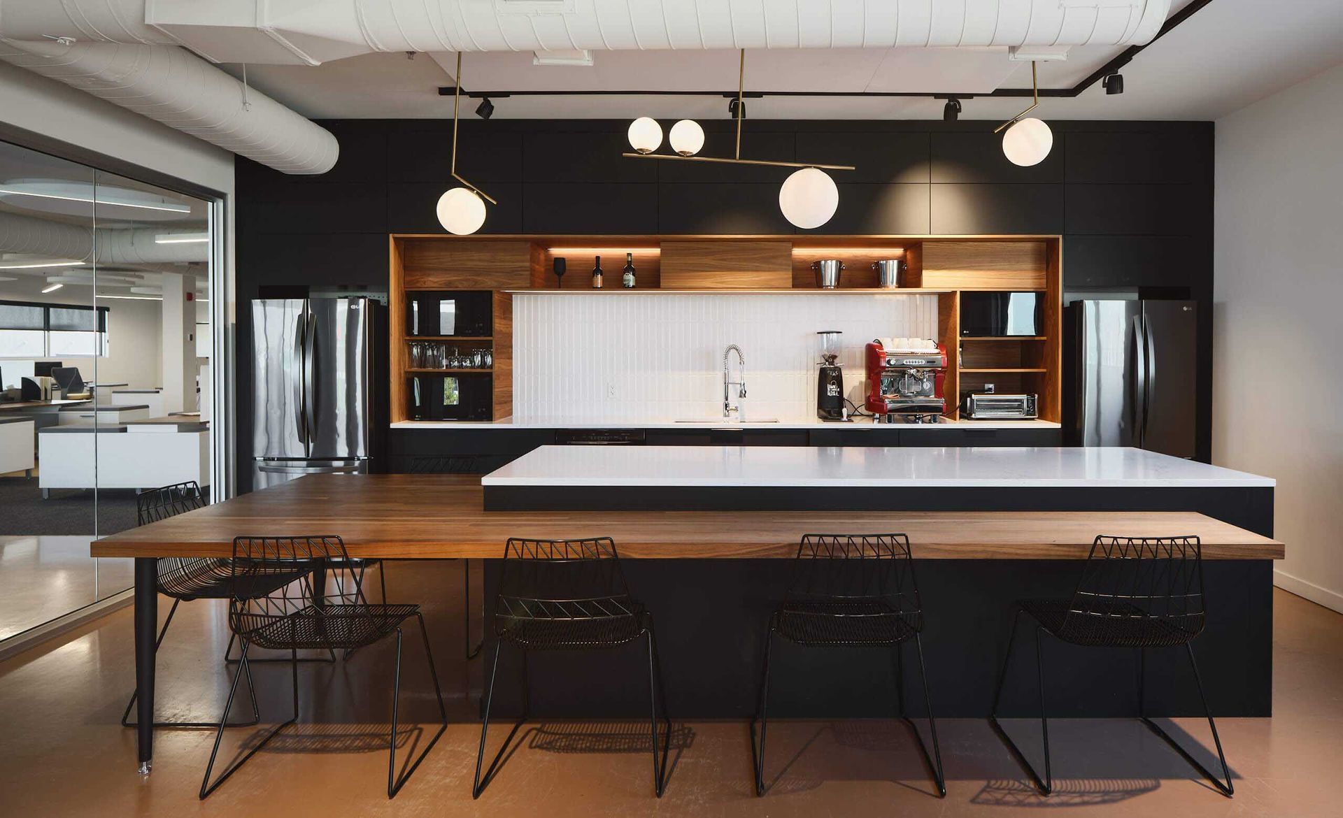 Cuisine de bureau avec placards fonc&eacute;s, plan de travail en bois, chaises noires et &eacute;clairage moderne.