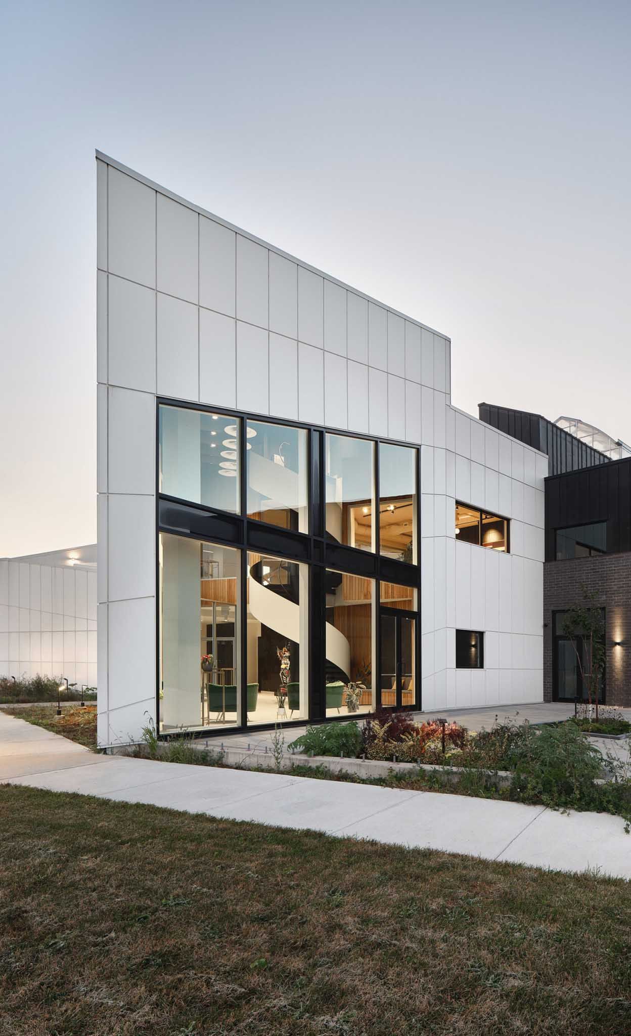B&acirc;timent blanc moderne avec de grandes baies vitr&eacute;es laissant appara&icirc;tre un escalier en colima&ccedil;on. Porte marron, touches de noir et petit jardin.