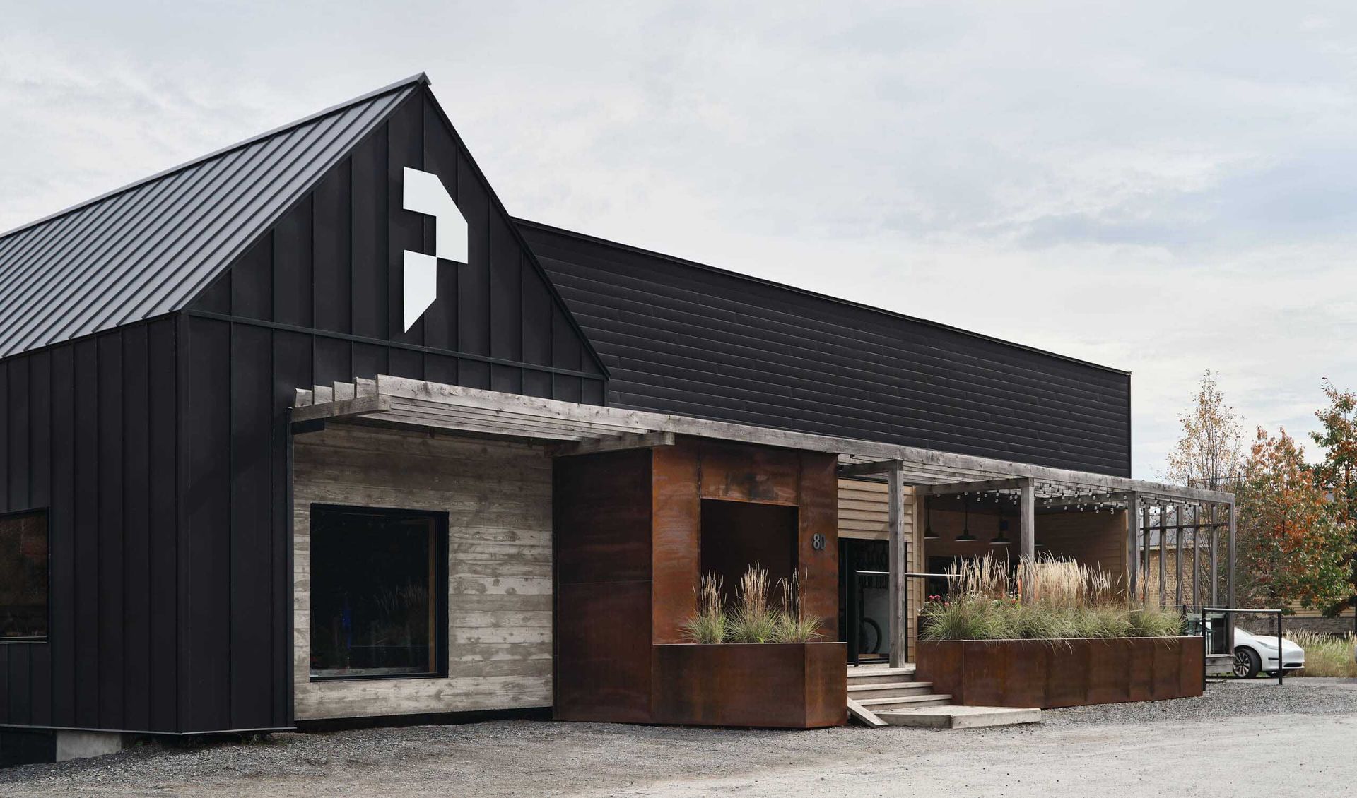 Bâtiment noir avec logo blanc ; éléments métalliques patinés, végétation et ciel nuageux.