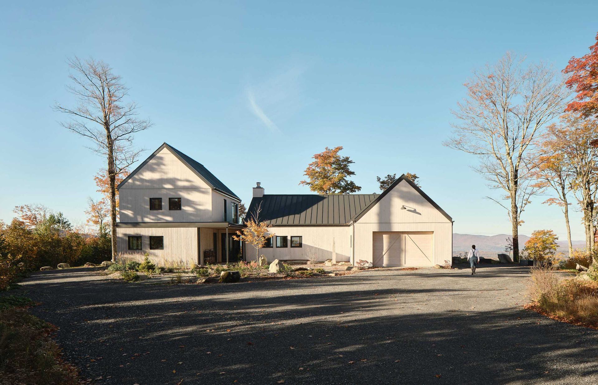 Maison moderne de couleur claire avec garage attenant, all&eacute;e en gravier et une personne qui s'en approche, par une journ&eacute;e ensoleill&eacute;e.