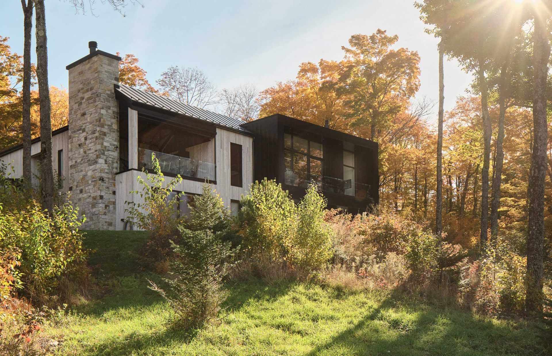 Maison moderne &agrave; flanc de colline, chemin&eacute;e en pierre, fa&ccedil;ade sombre, entour&eacute;e d'arbres aux couleurs d'automne.