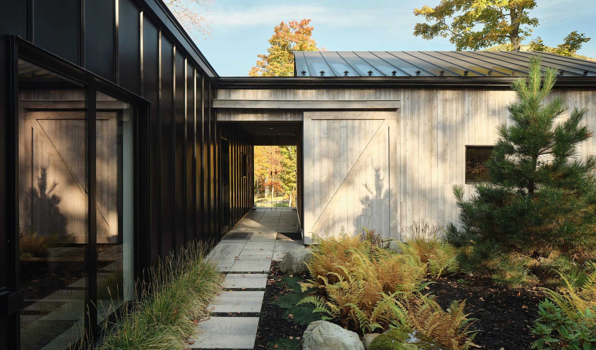 Extérieur d'un bâtiment moderne avec bardage en bois patiné, bardage en métal foncé, chemin de pierre et verdure.