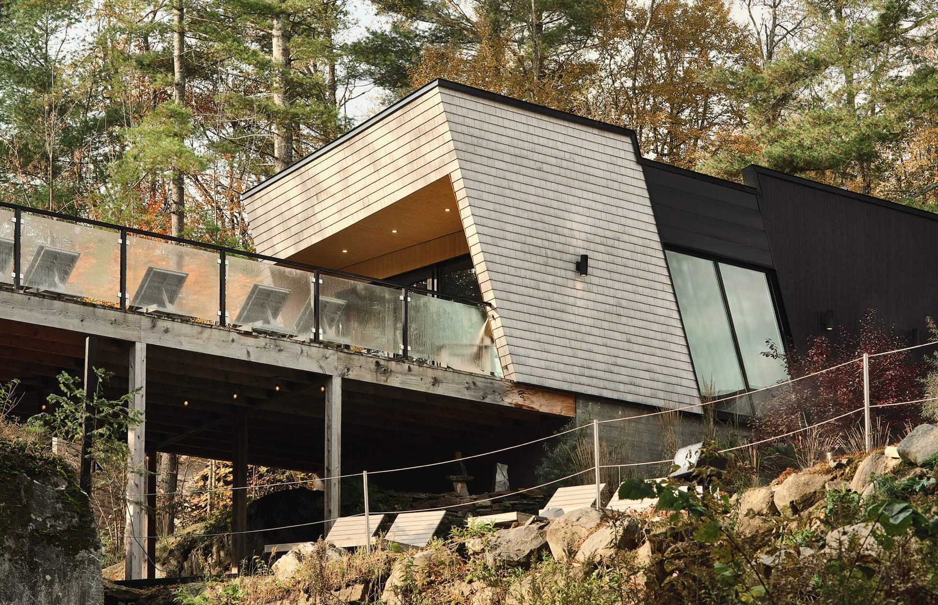 Maison moderne avec toit en pente, terrasse en bois et grandes fen&ecirc;tres, situ&eacute;e dans une zone bois&eacute;e.