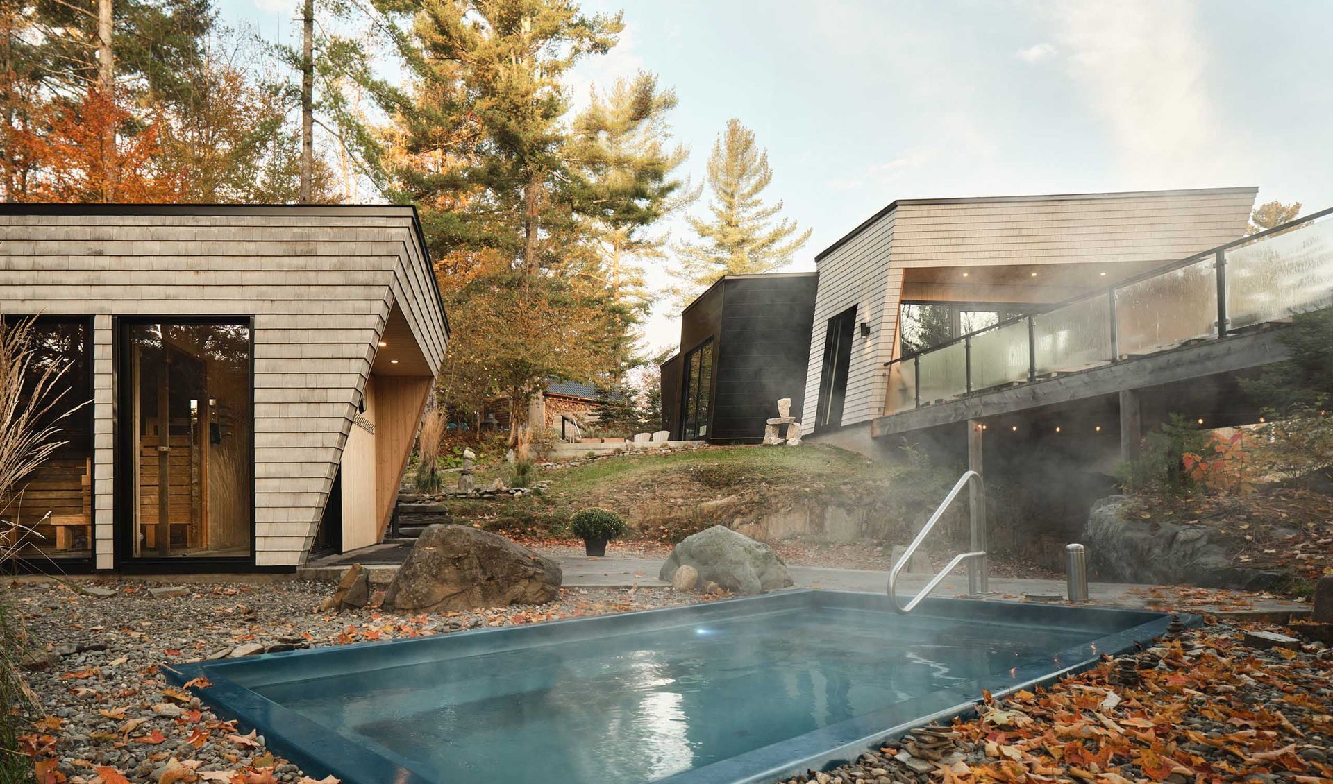 Spa extérieur avec piscine rectangulaire, vapeur s'échappant. Cabanes modernes à bardage bois. Feuillage d'automne visible.