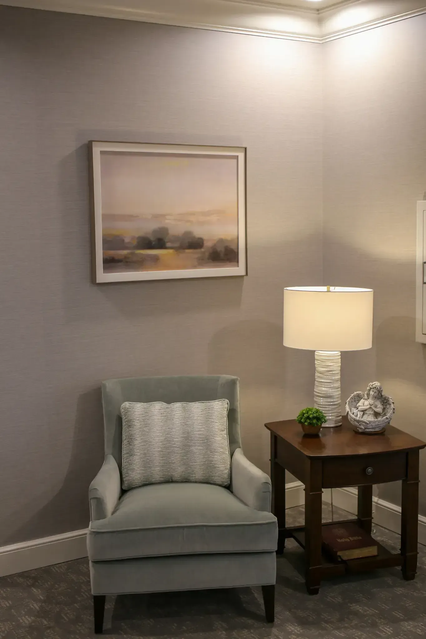 Cozy corner with gray chair, lamp on side table, and framed landscape in a softly lit room