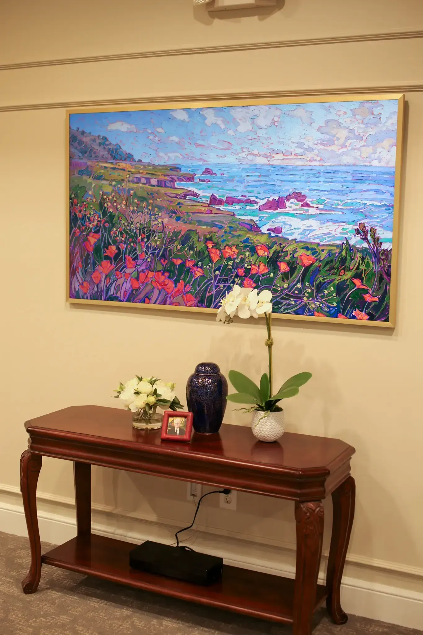 Framed coastal painting above a wooden console table with flowers and decor in a beige hallway