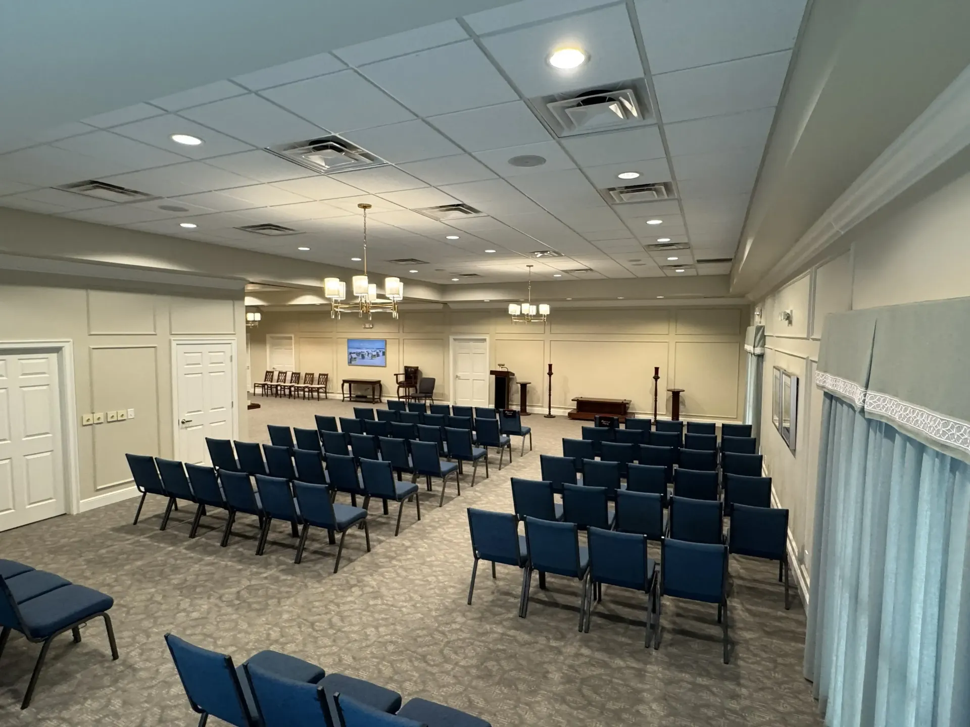 Empty room with rows of blue chairs, ceiling lights, and a small stage at the front.