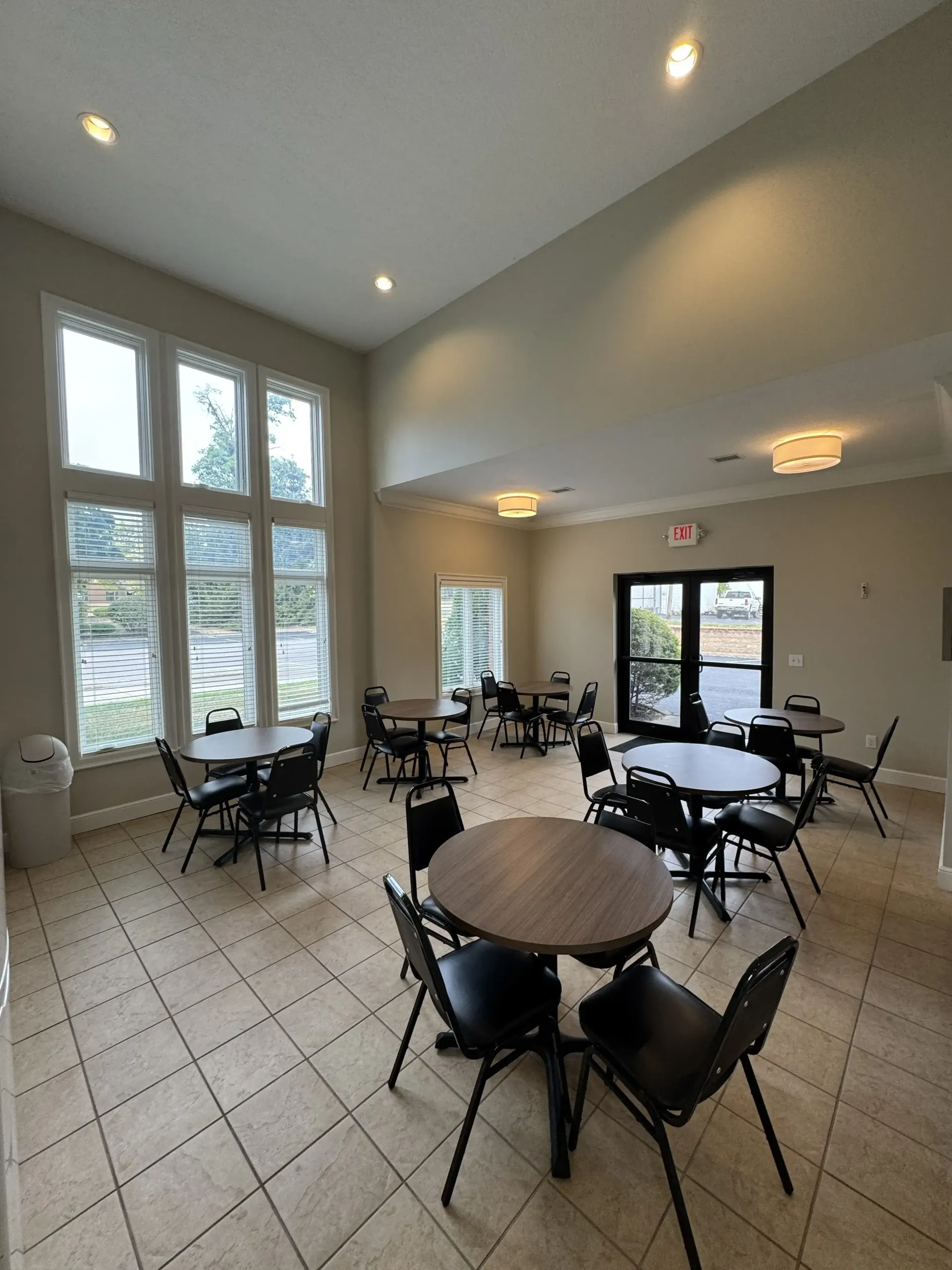 Bright hall with round tables, black chairs, large windows, tiled floor, and wall sconces.