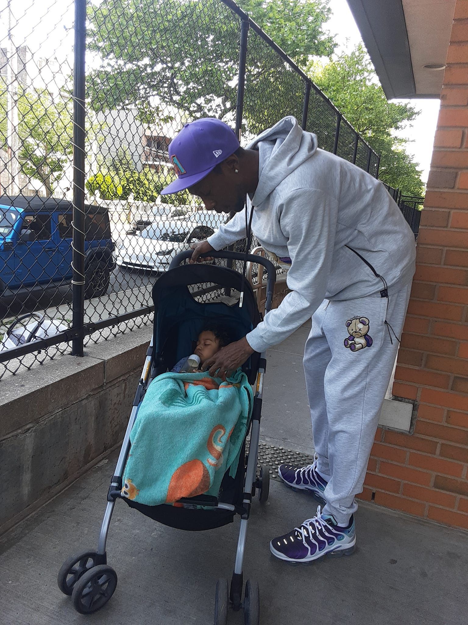 Adult in white tracksuit adjusts a stroller on a sidewalk beside a brick wall and chain-link fence.