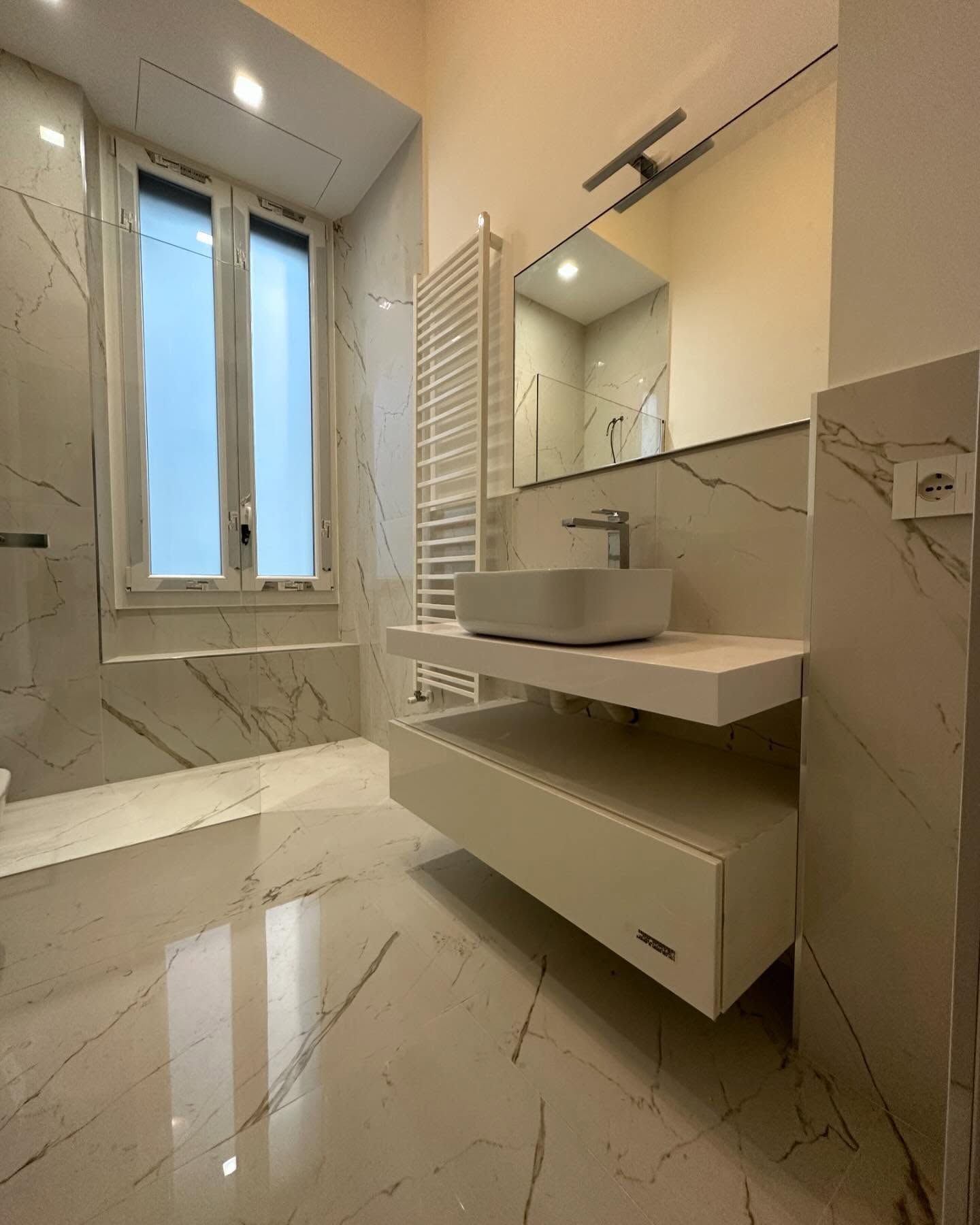 Bagno moderno con piastrelle in marmo, lavabo sospeso, specchio e finestra che lascia entrare la luce naturale.