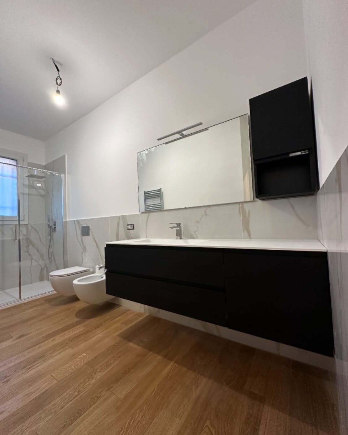 Bagno moderno con mobile lavabo nero, ampio specchio, piastrelle bianche, doccia in vetro, WC e pavimento effetto legno.