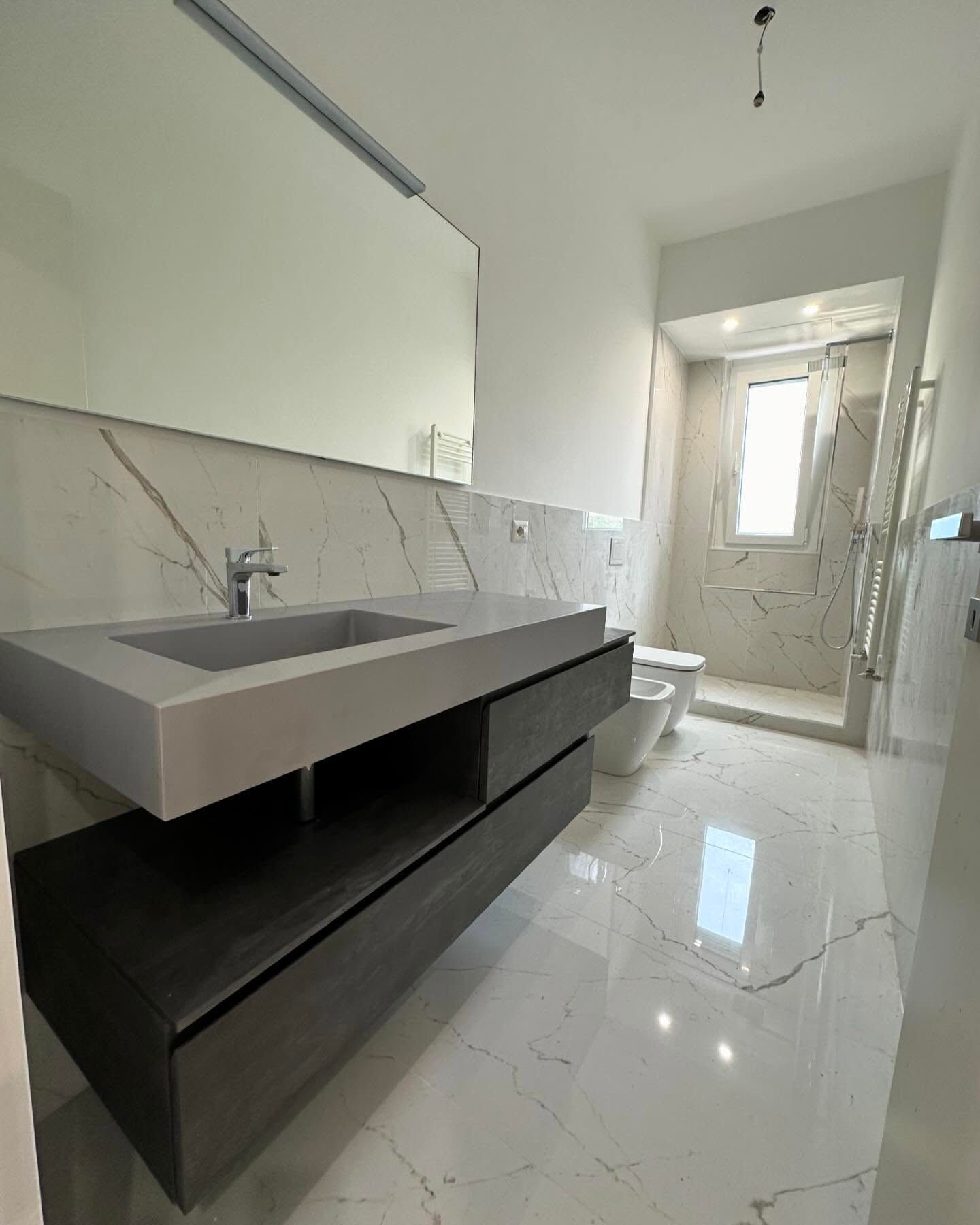 Bagno moderno con mobile lavabo sospeso, ampio specchio, piastrelle in marmo bianco e doccia in fondo.