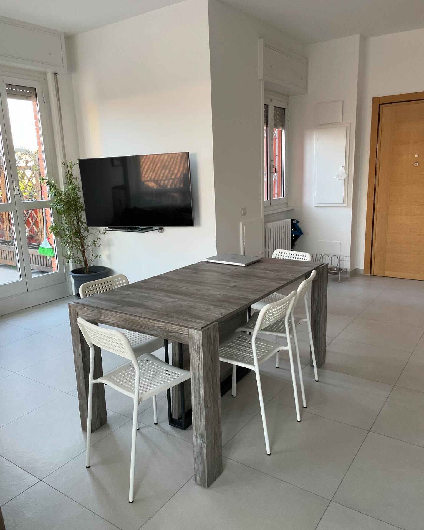 Zona pranzo moderna con tavolo in legno, sedie bianche, TV a parete e pavimento piastrellato.