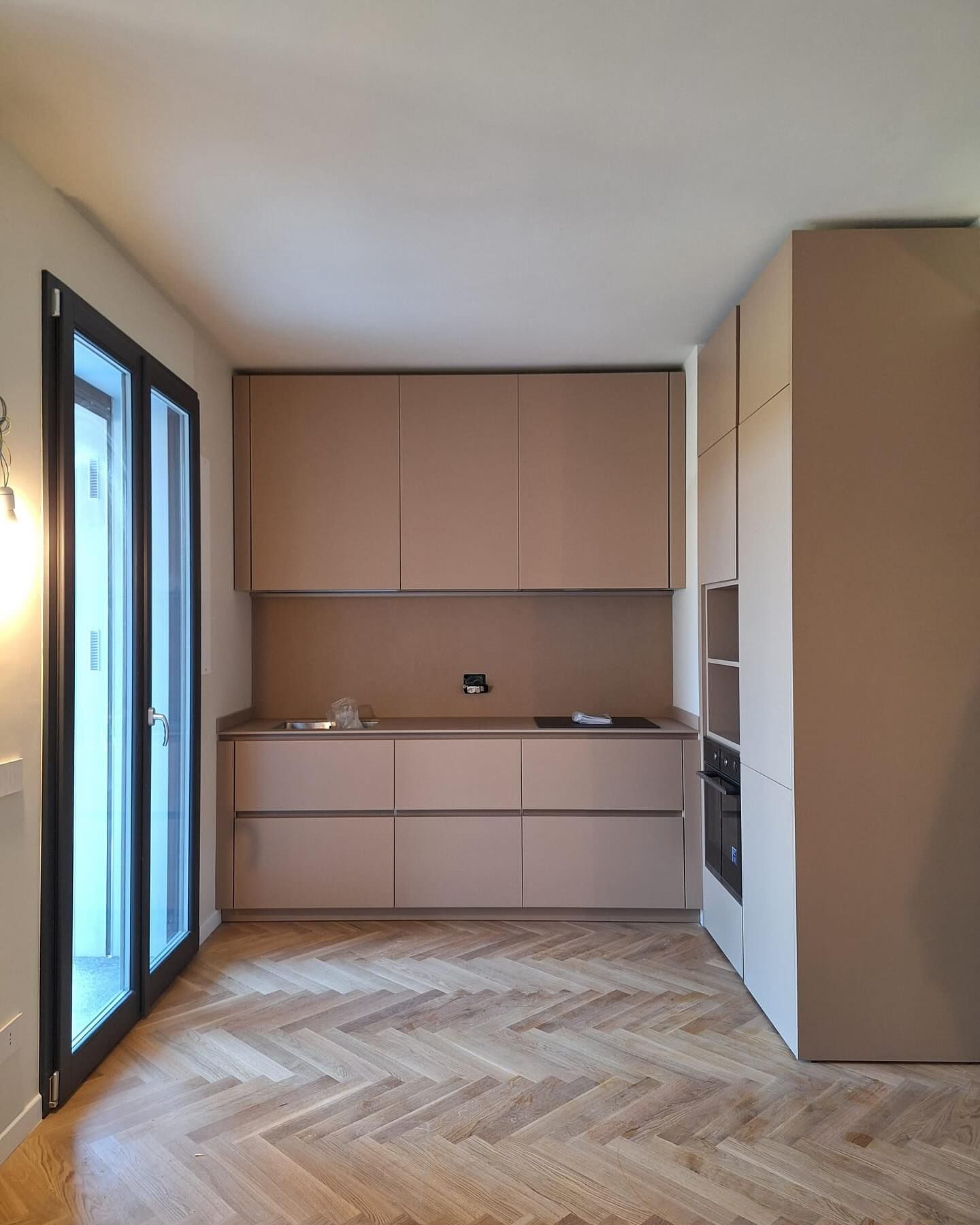 Cucina moderna con mobili beige, pavimento in parquet a spina di pesce e ampie porte a vetri sulla sinistra.