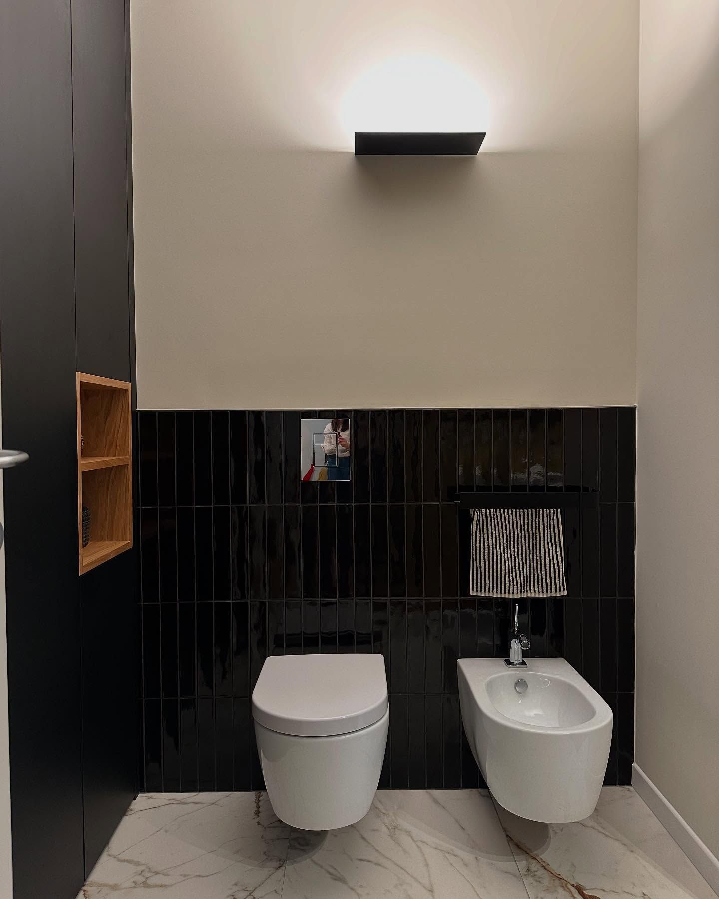 Bagno moderno con rivestimento a piastrelle nere, WC sospeso, lavabo bianco e lampada a soffitto.