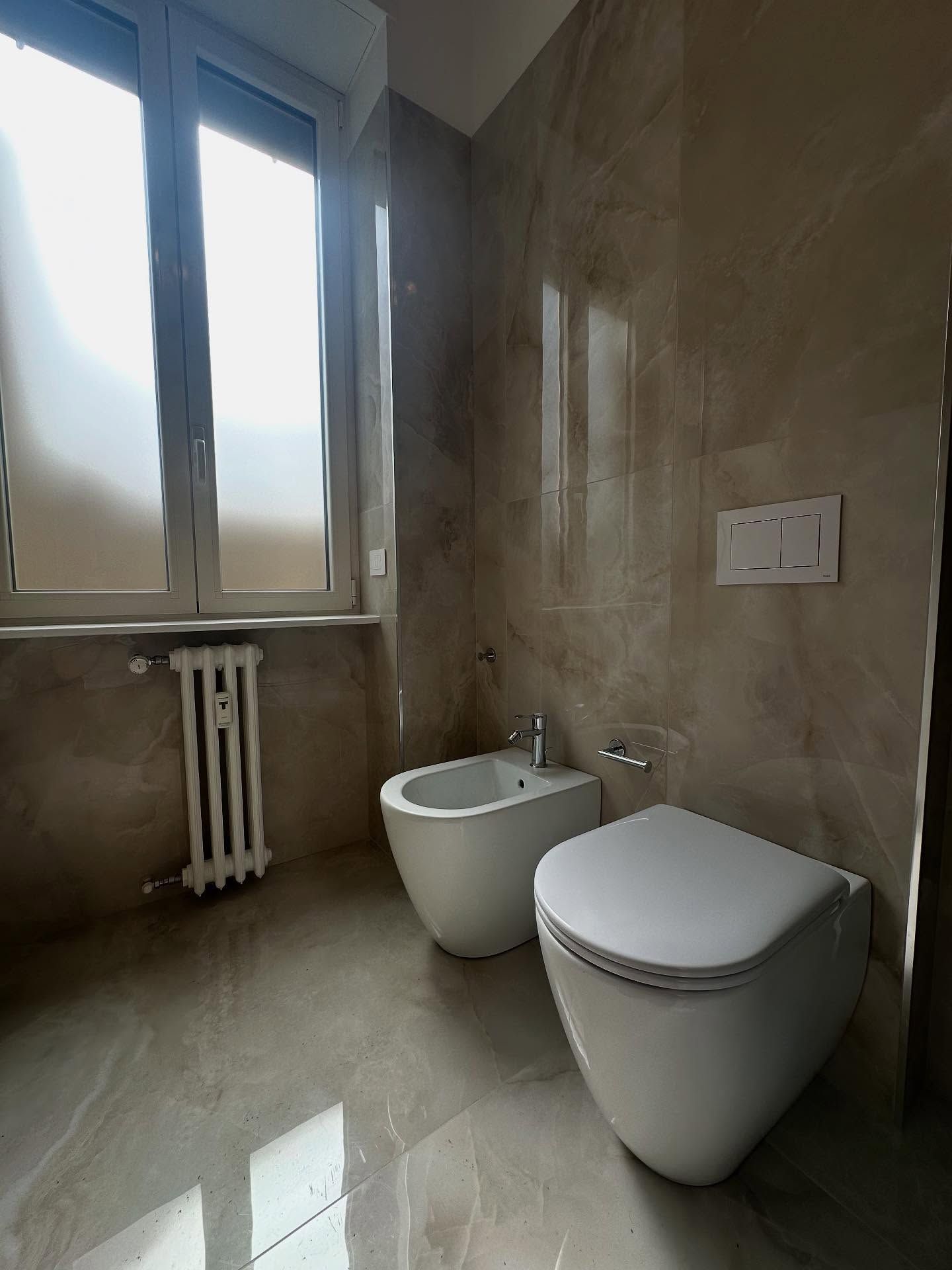 Bagno moderno con pareti rivestite di piastrelle beige, finestra, termosifone, lavabo e WC.