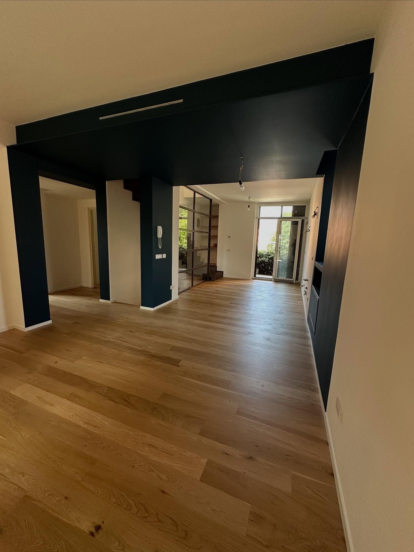 Corridoio interno moderno con pavimento in legno, pareti scure e finestre luminose in fondo.