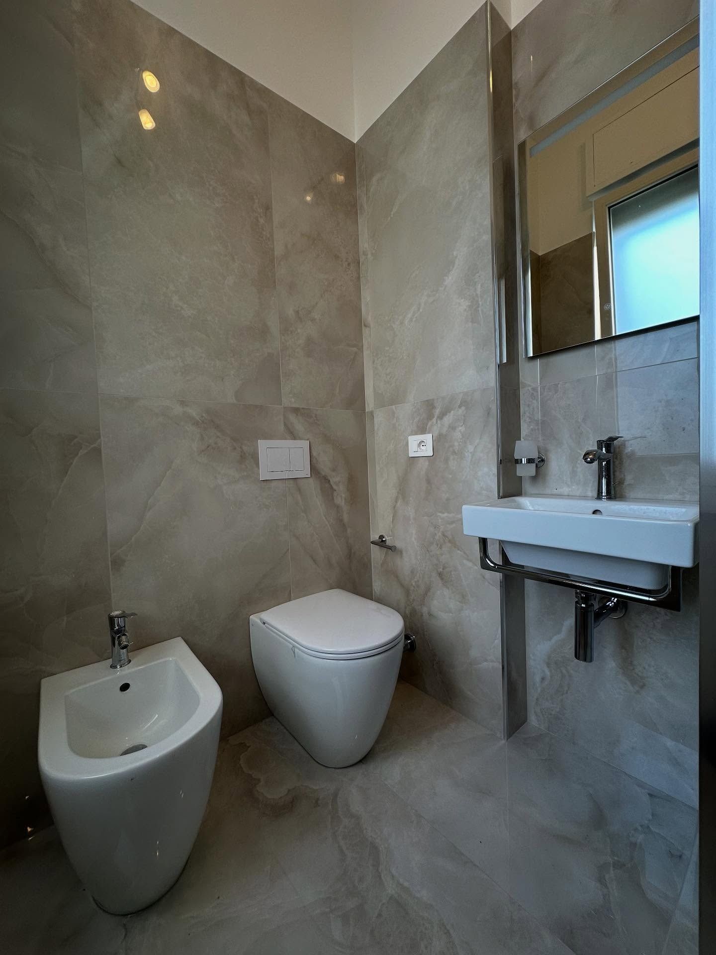 Bagno moderno con WC, bidet, lavabo a parete e specchio rivestito in piastrelle di pietra grigia.