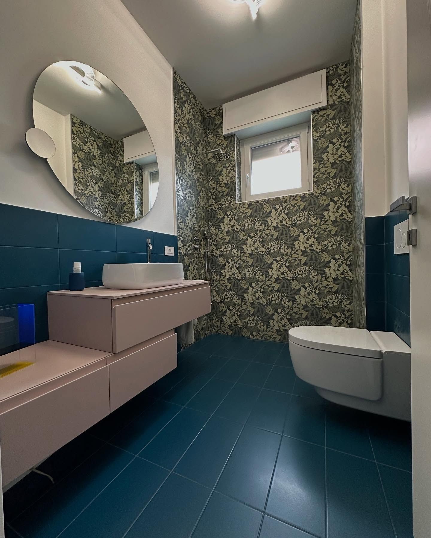 Bagno compatto con pavimento in piastrelle color verde acqua, lavabo a parete, specchio rotondo e carta da parati floreale.