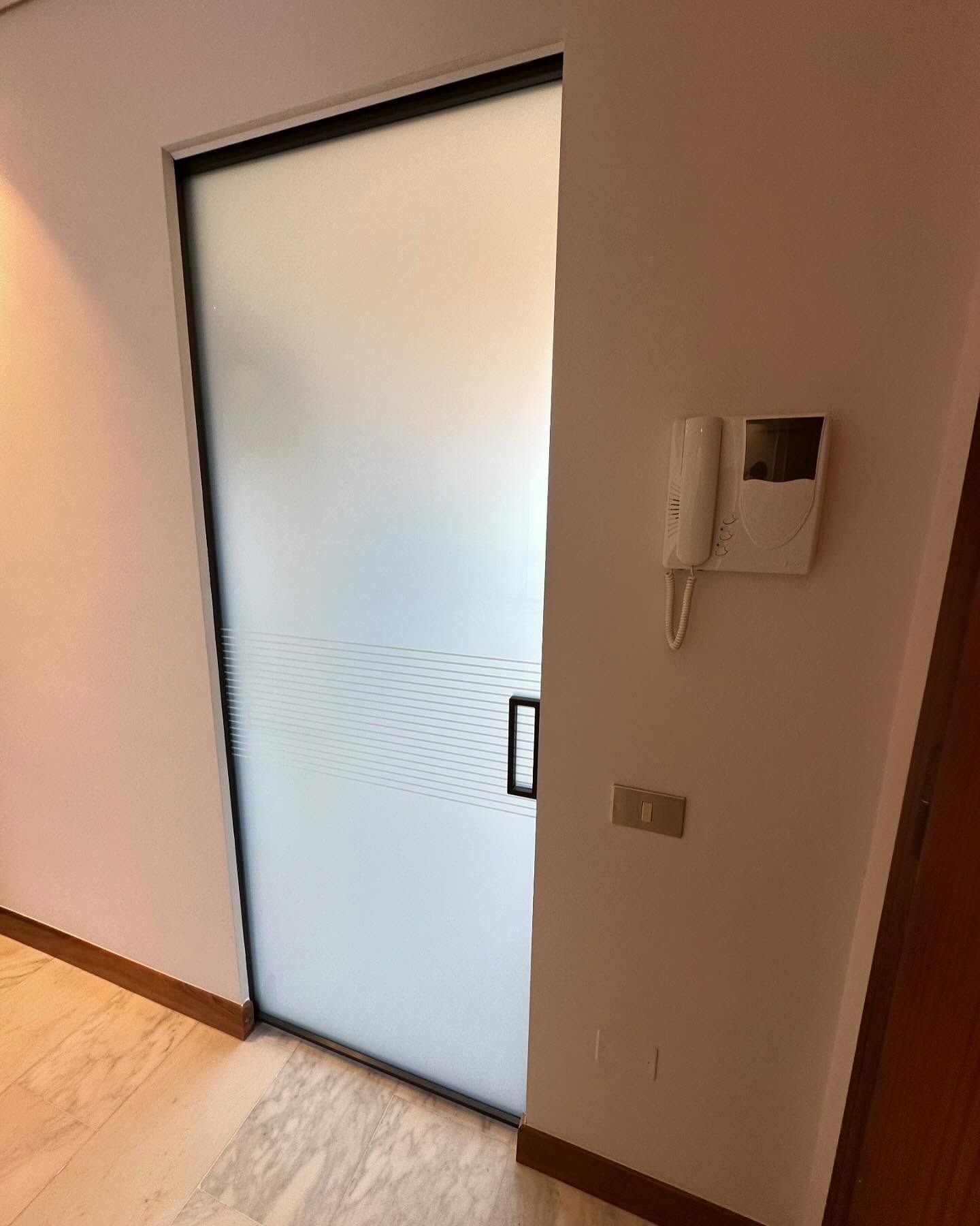 Porta in vetro satinato in un corridoio con citofono a parete e pavimento in legno chiaro.