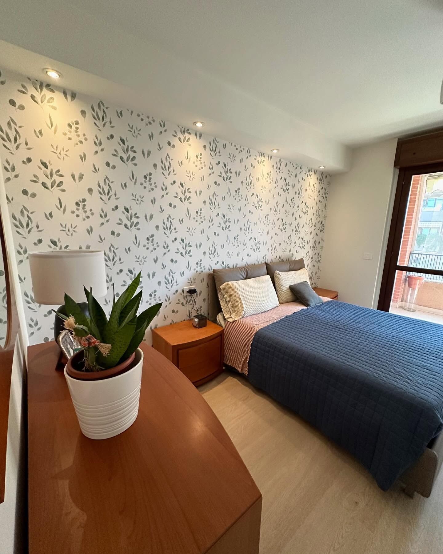 Camera da letto luminosa con carta da parati a motivi geometrici, letto, comò in legno e pianta in vaso vicino alla finestra.