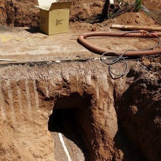 Excavation site with a dark tunnel entrance, hoses, and a small control box on the dirt surface.