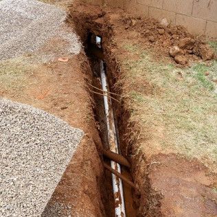 Broken ground exposing a rusted pipe in a narrow trench beside a wall.