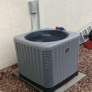 Gray outdoor air conditioner unit beside a house wall on gravel, with red and blue valves on top.