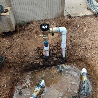 Outdoor plumbing pipes and valves beside a dirt patch and concrete slab