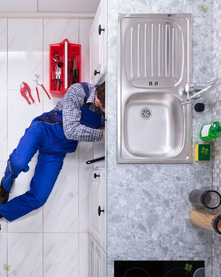 Person in blue overalls working on a kitchen cabinet beside a sink and red tool kit.