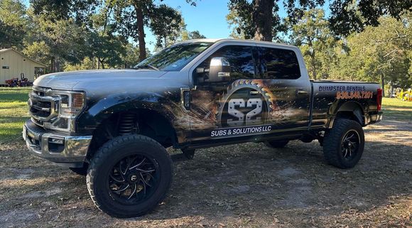 Silver lifted pickup truck with black wheels, custom wrap on side, parked outside.