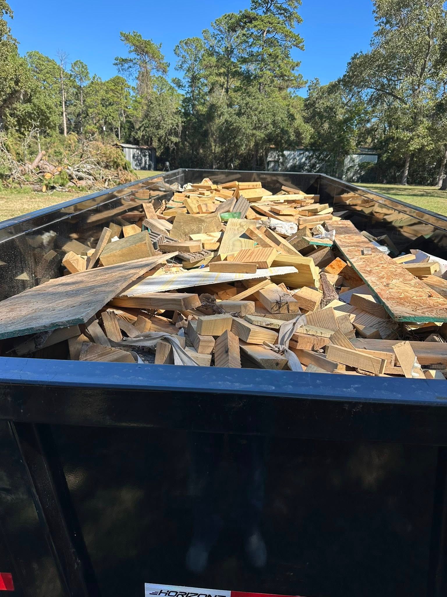 Black dumpster filled with construction debris, outdoors under a blue sky and trees.