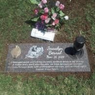 Gravestone with flowers and a black candle in a grassy cemetery plot.