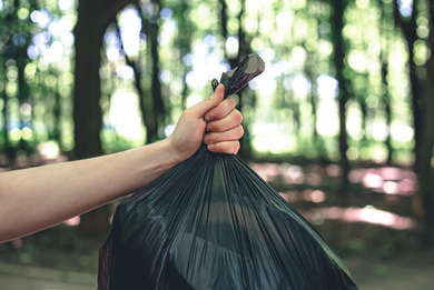 Mano sosteniendo una bolsa de basura negra contra un fondo de bosque borroso.