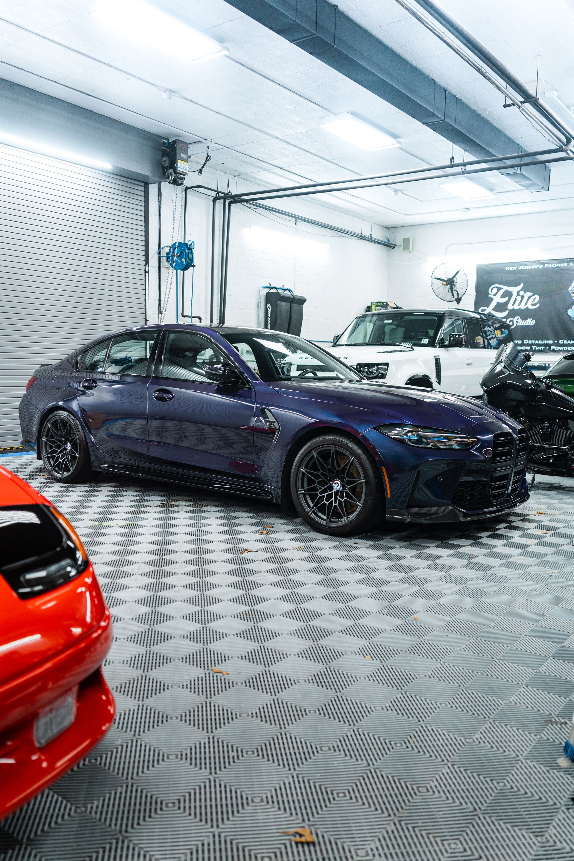 Dark purple sports sedan in a bright car garage, with a red car partially visible in the foreground.