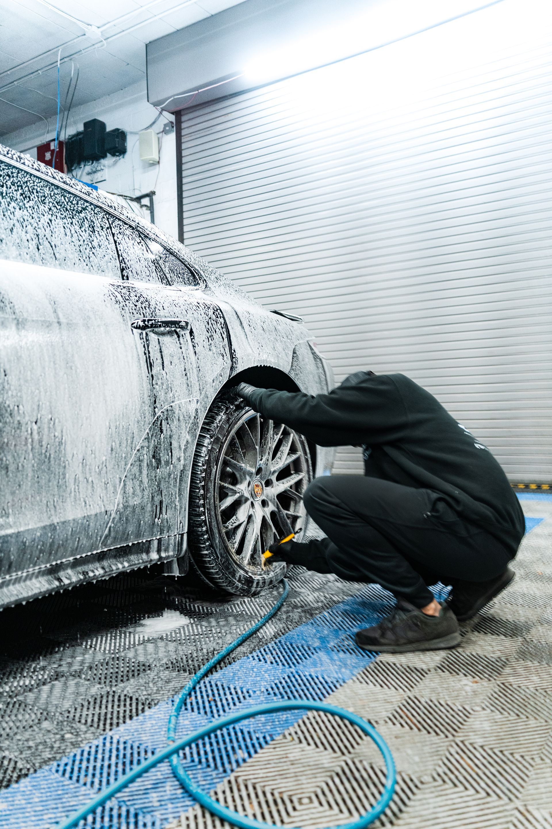 Person washing a white car wheel in a garage, crouched beside a foam-covered vehicle.