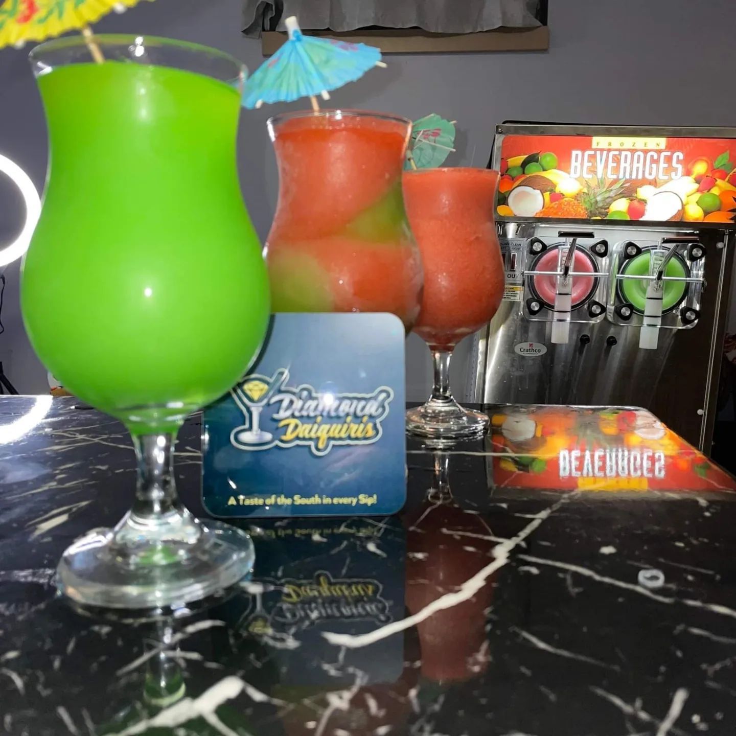 Three frozen cocktails in glass cups with mini umbrellas stand on a marble bar near a frozen beverage machine.