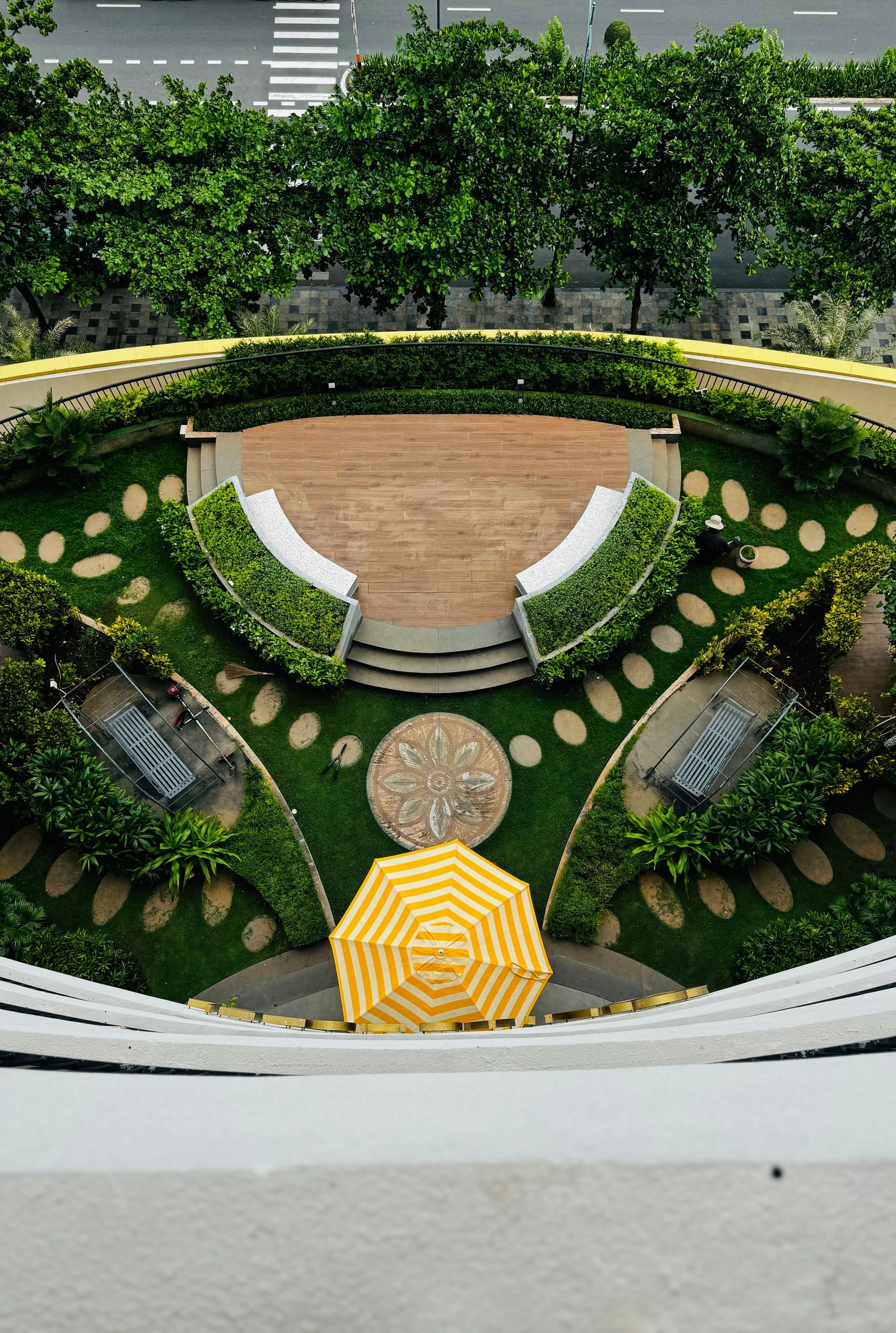 Circular landscaped garden with white benches, green shrubs, and a yellow umbrella in the center