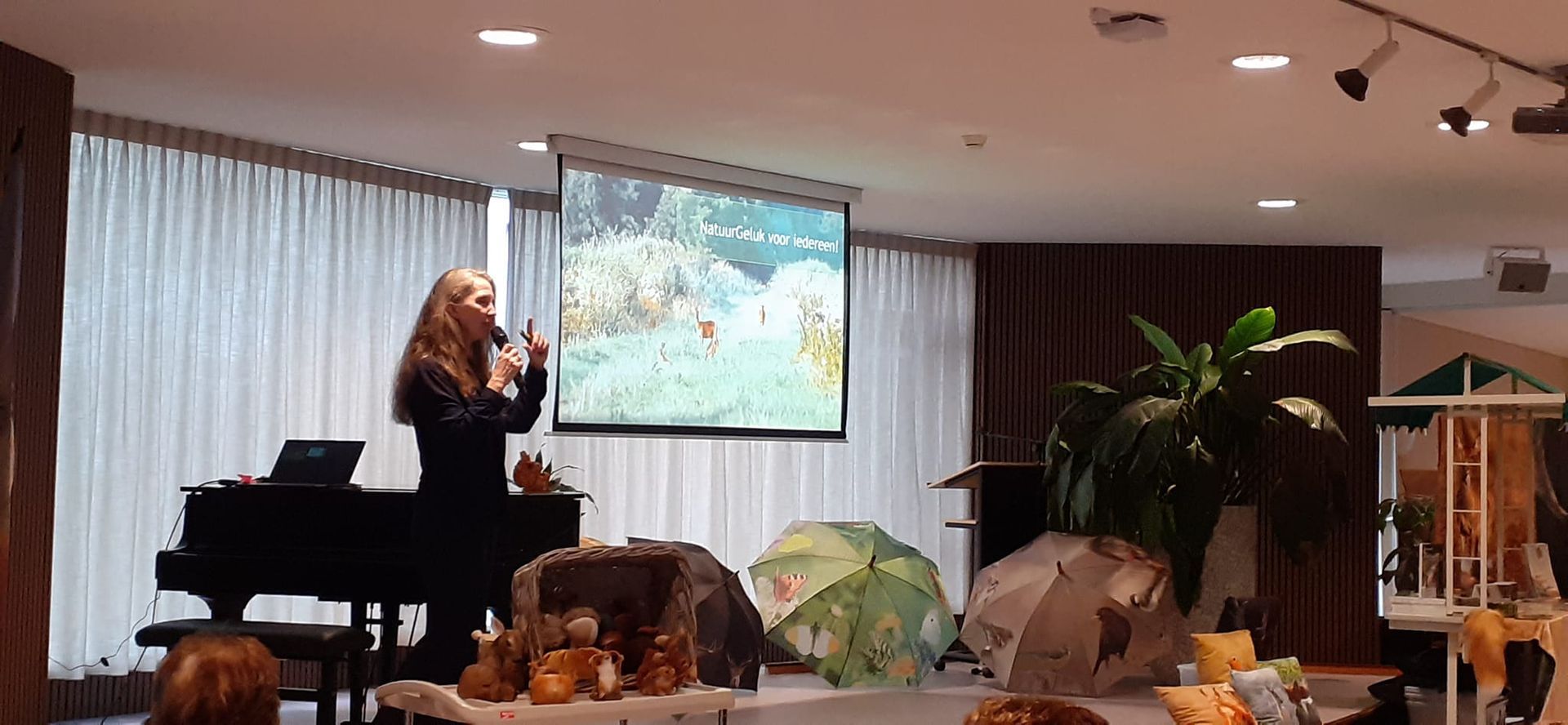 Een vrouw presenteert met een microfoon, een scherm waarop een natuurscène te zien is 