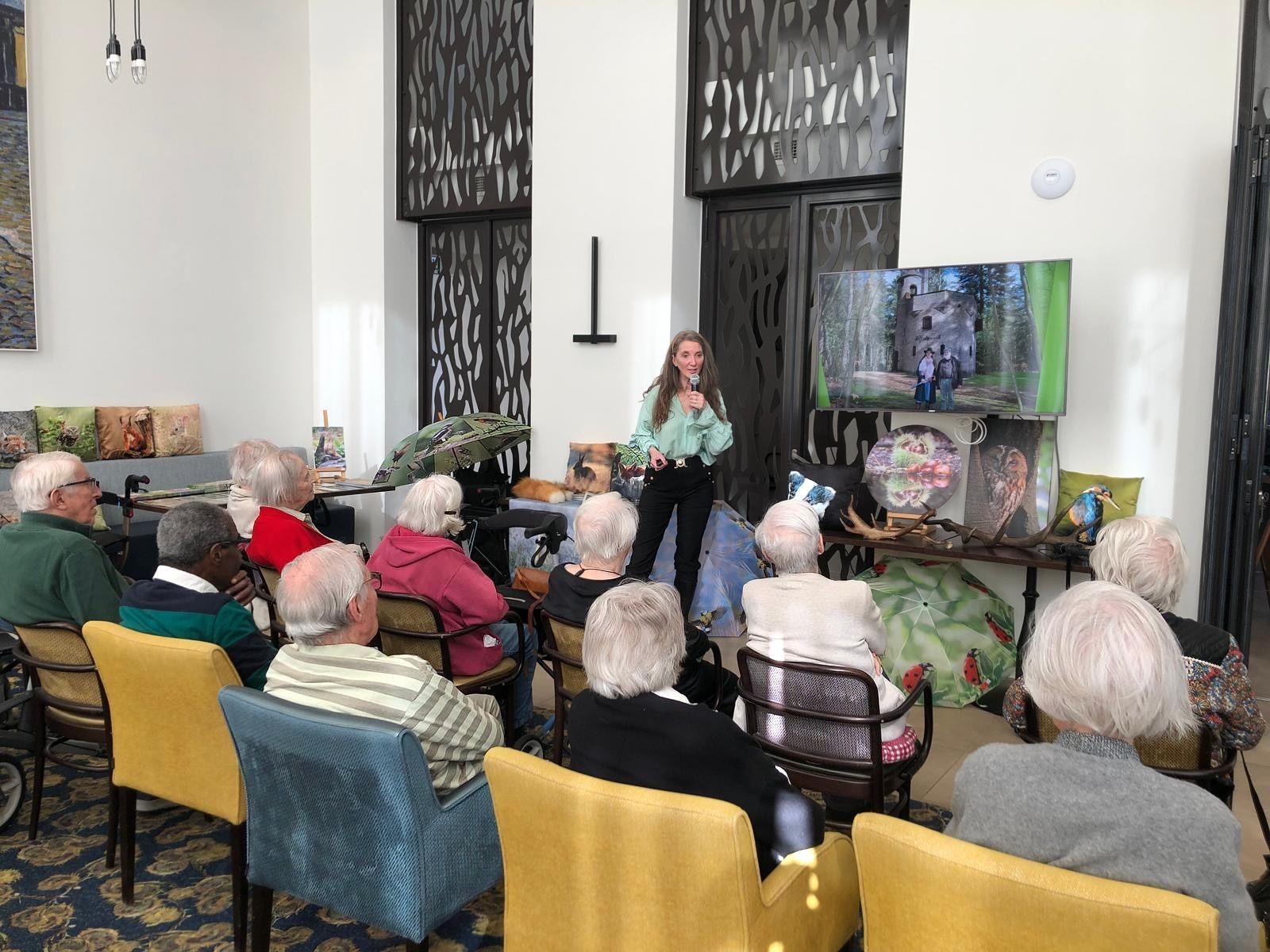 Een vrouw geeft een presentatie aan een zittend publiek in een lichte ruimte. Op een scherm wordt een video weergegeven.