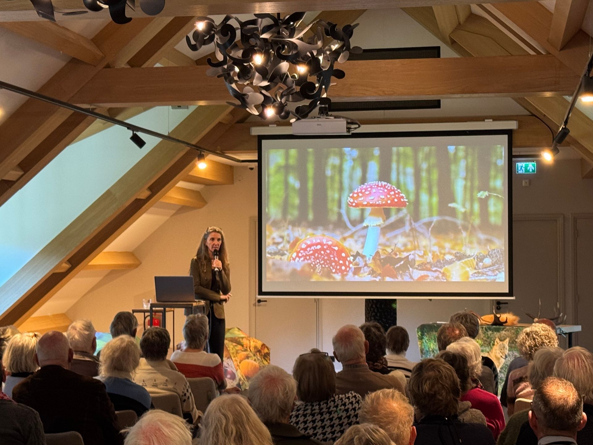 Vrouw geeft een presentatie over paddenstoelen aan een publiek; geprojecteerd beeld van paddenstoelen.