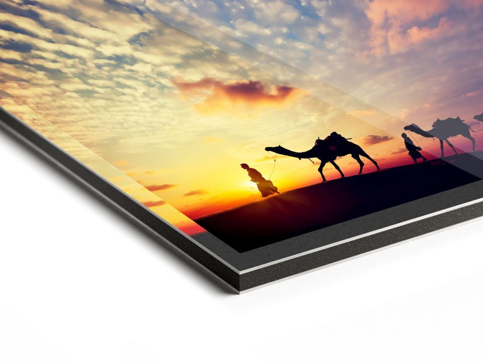 Silhouetten van kamelen die bij zonsondergang de woestijn doorkruisen, gemonteerd op een zwarte metalen print. Oranje en blauwe lucht.