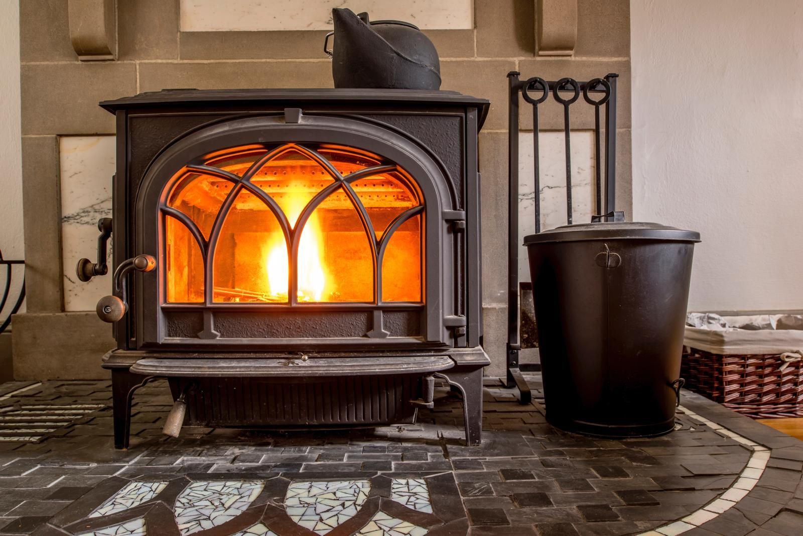 Wood-burning stove with flames inside, next to a metal bucket and fireplace tools. The stove is on a patterned tile floor.