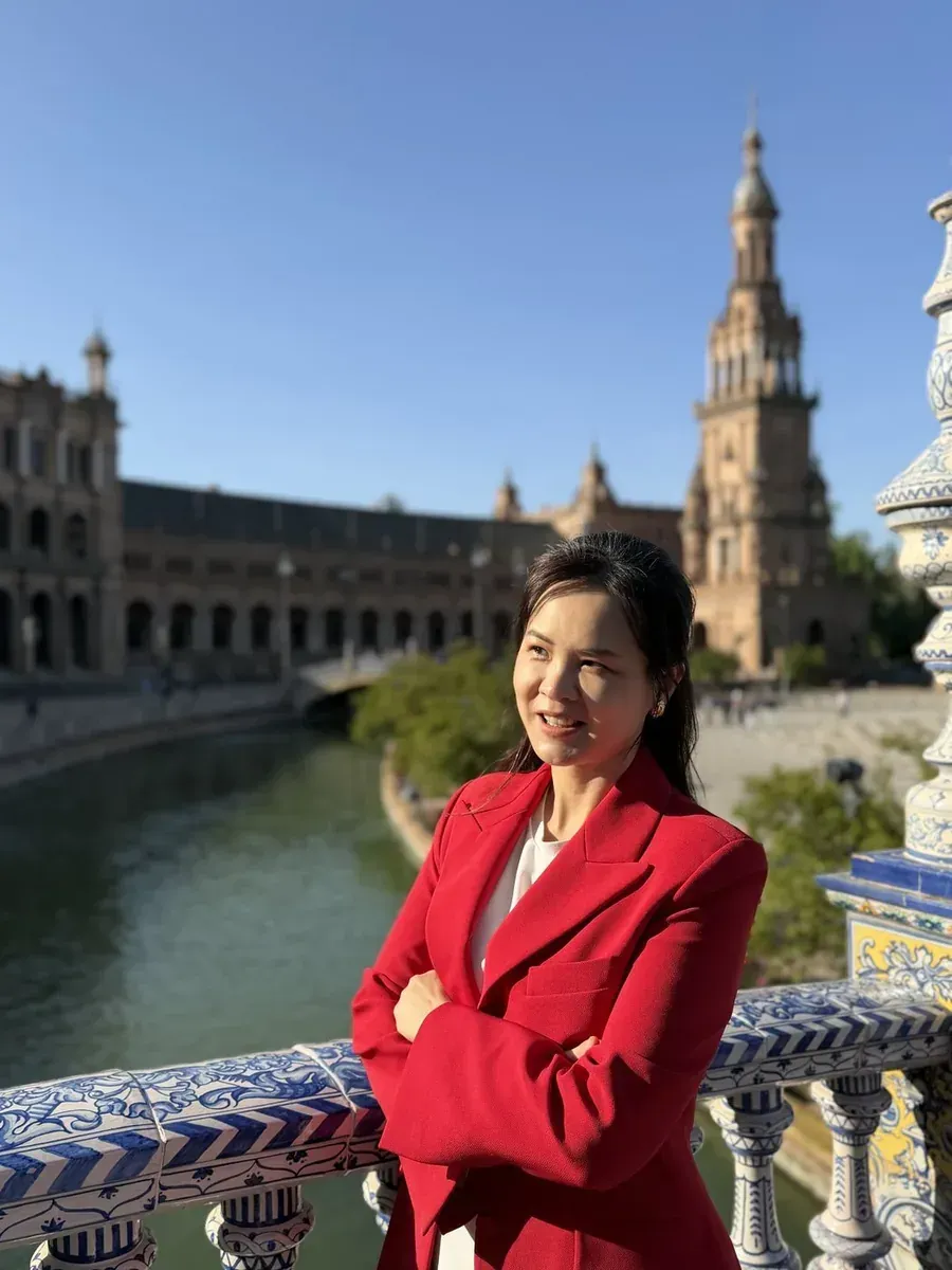 CP 穿紅外套站在塞維亞的西班牙廣場，雙手交叉看向遠方