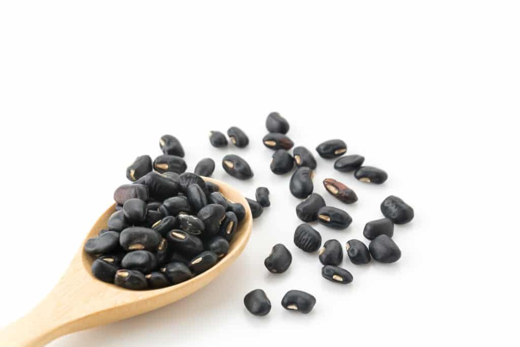 Black beans in a wooden spoon, spilling onto a white surface.